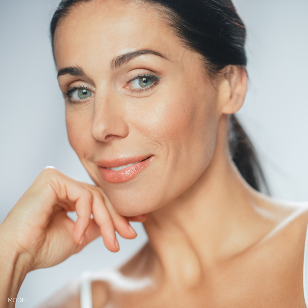 close up of woman with dark hair looking at camera, with hand leaning on chin